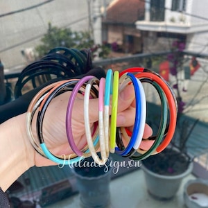 Könnte beinhalten: Eine Sammlung bunter Armreifen mit schwarzen und weißen Akzenten. Die Armreifen sind aus einem glatten, glänzenden Material gefertigt und in einer Hand übereinander gestapelt.