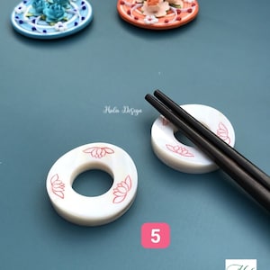 May include: White ceramic chopstick rests with a red lotus flower design. The rests are stacked on top of each other. Two black chopsticks are laying on top of the rests. The background is a light blue color.