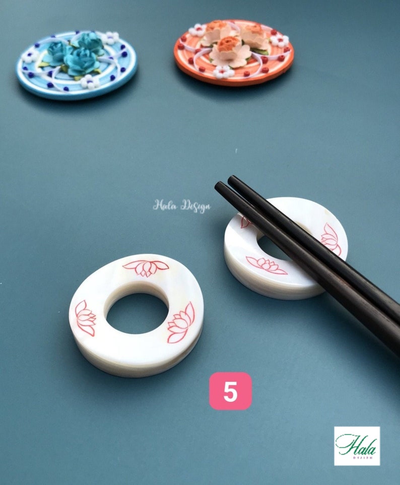 May include: White ceramic chopstick rests with a red lotus flower design. The rests are stacked on top of each other. Two black chopsticks are laying on top of the rests. The background is a light blue color.