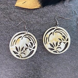 May include: A pair of cream-colored wooden earrings with a delicate floral design. The earrings are circular and have a silver hook.