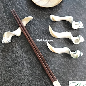Peut inclure: Baguettes chinoises marron foncé avec des embouts blancs reposent sur un repose-baguettes blanc et beige. Trois autres supports sont disposés à proximité. Un bol en bois est en arrière-plan.