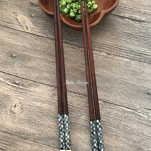 May include: Set of four brown wooden chopsticks with decorative mother-of-pearl inlays. The chopsticks are arranged on a wooden surface with a bowl of green peas in the background.