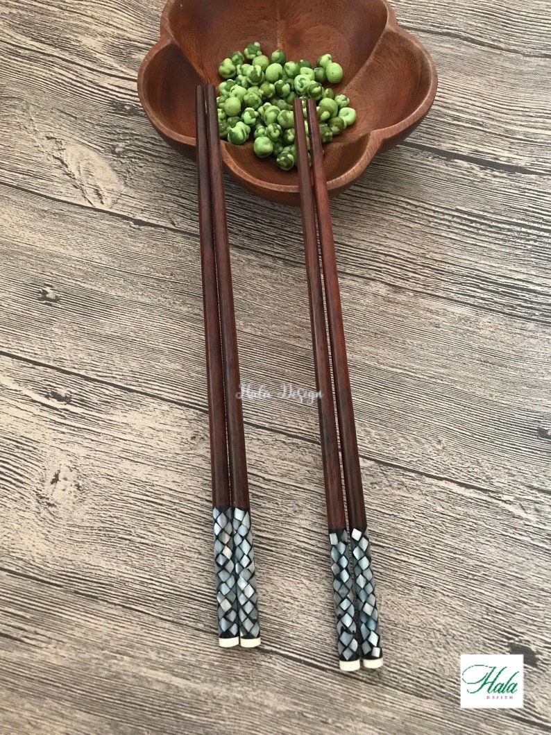 May include: Set of four brown wooden chopsticks with decorative mother-of-pearl inlays. The chopsticks are arranged on a wooden surface with a bowl of green peas in the background.
