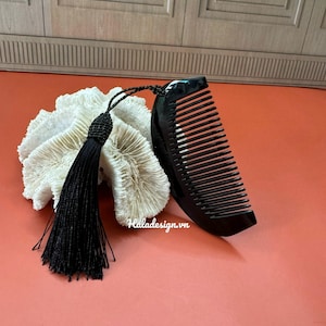 Puede incluir: Un peine negro con dientes finos y un mango curvo descansa junto a un objeto blanco similar al coral. Una borla negra está unida al peine. El fondo es de color naranja liso.