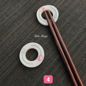 May include: Two white ceramic chopstick rests with a green and red lotus flower design. The rests are on a dark gray textured surface. Two brown wooden chopsticks are resting on the rests.