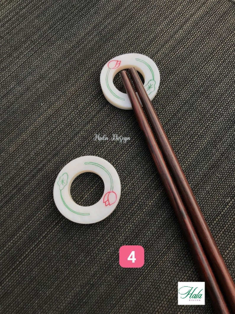 May include: Two white ceramic chopstick rests with a green and red lotus flower design. The rests are on a dark gray textured surface. Two brown wooden chopsticks are resting on the rests.