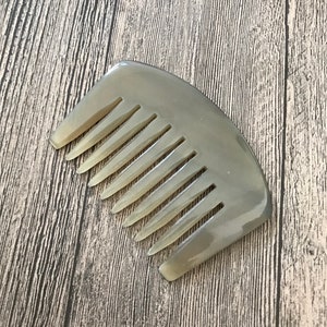 K&ouml;nnte beinhalten: Ein hellbrauner Kamm aus Horn mit abgerundeter Form und einer Reihe von Z&auml;hnen. Der Kamm liegt auf einer Holzoberfl&auml;che.