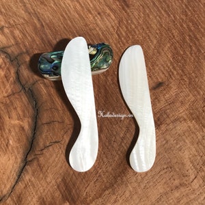 May include: Two white mother of pearl butter knives with a curved handle. The knives are on a wooden surface.