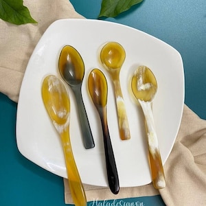 Puede incluir: Cinco cucharas hechas de cuerno pulido, de color marrón claro, están dispuestas en un plato blanco. Las cucharas son de diferentes tamaños y formas, y tienen un aspecto natural y orgánico.