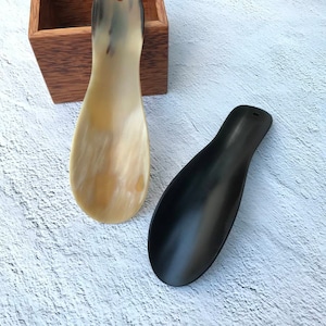May include: Two shoe horns, one made of black plastic and one made of natural horn. The horn shoe horn is resting on a wooden box.