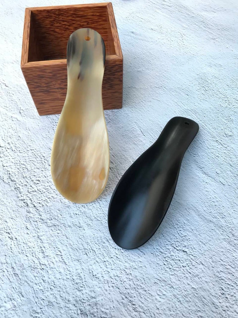 May include: Two shoe horns, one made of black plastic and one made of natural horn. The horn shoe horn is resting on a wooden box.