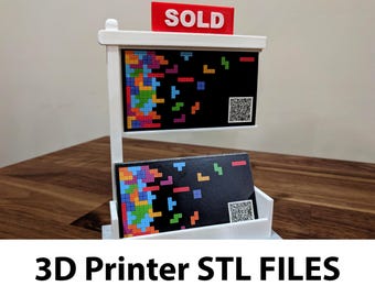 May include: A white 3D printed stand with a sign that says "SOLD" and two black cards with a colorful Tetris pattern. The stand is on a wooden surface.