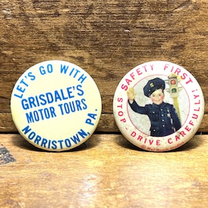 May include: Two vintage pinback buttons. One is cream-colored with blue text that reads "Let's Go With Grisdale's Motor Tours Norristown, PA." The other button has a child in a police uniform with the text "Safety First! Drive Carefully!"