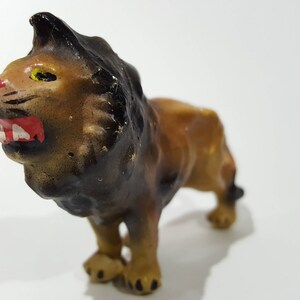 May include: A small, vintage lion figurine with a brown body, black mane, and a red mouth. The lion is posed in a roaring stance, with its mouth open, revealing white teeth. The figurine is made of a hard material and has a slightly worn appearance.