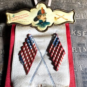 May include: Vintage firefighter's badge with a portrait, ladder, and trumpet illustrations. Below, two diamond-shaped patriotic ornaments with red and white stripes and blue accents hang from a red ribbon.