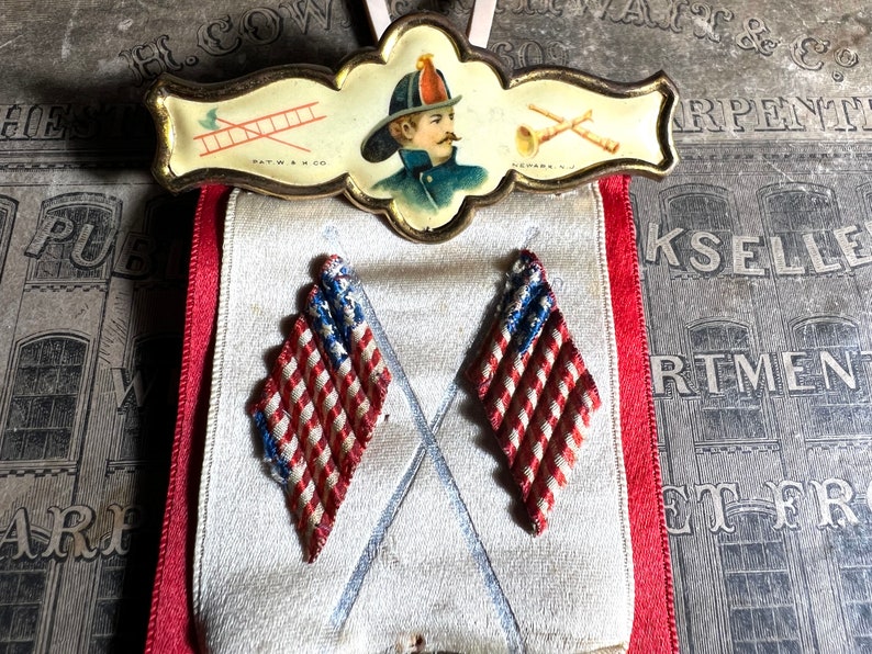 May include: Vintage firefighter's badge with a portrait, ladder, and trumpet illustrations. Below, two diamond-shaped patriotic ornaments with red and white stripes and blue accents hang from a red ribbon.