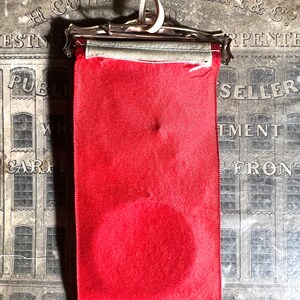 May include: A vintage red ribbon with a silver metal clasp and a cream-colored ribbon for hanging. The ribbon has a circular indentation and text at the bottom. The background features an old advertisement.