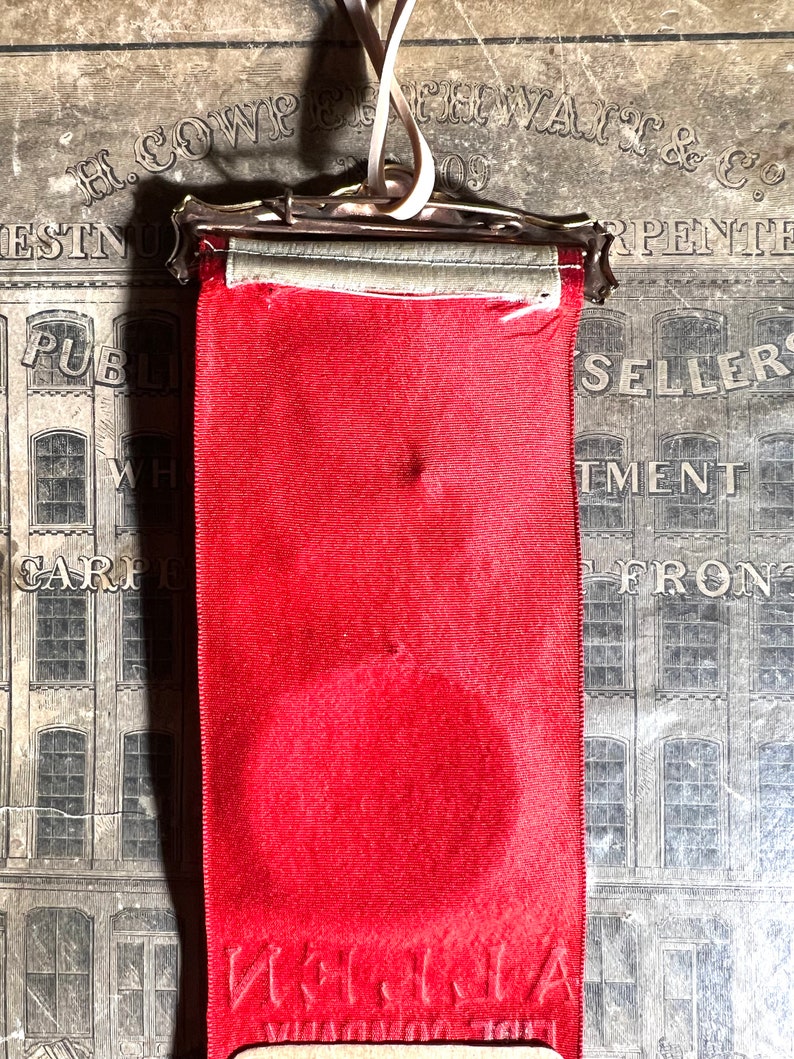 May include: A vintage red ribbon with a silver metal clasp and a cream-colored ribbon for hanging. The ribbon has a circular indentation and text at the bottom. The background features an old advertisement.