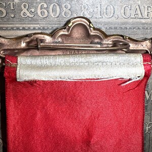 May include: A vintage award ribbon with a red fabric banner and a cream-colored top section. The metal pin reads "THE WHITEHEAD & HOAG, NEWARK, N.J." in embossed lettering. The ribbon is attached to a decorative metal pin.