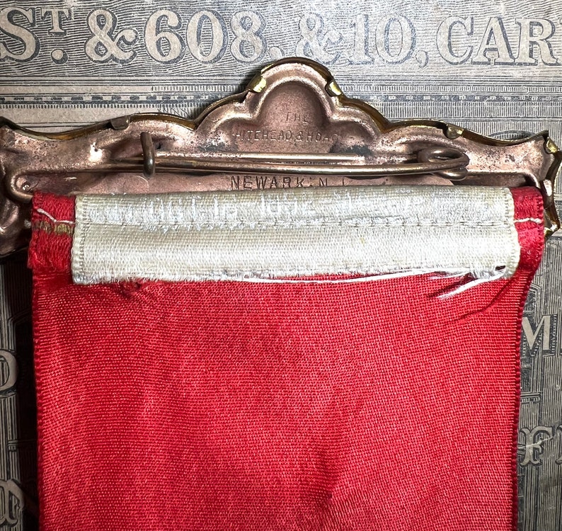 May include: A vintage award ribbon with a red fabric banner and a cream-colored top section. The metal pin reads "THE WHITEHEAD & HOAG, NEWARK, N.J." in embossed lettering. The ribbon is attached to a decorative metal pin.