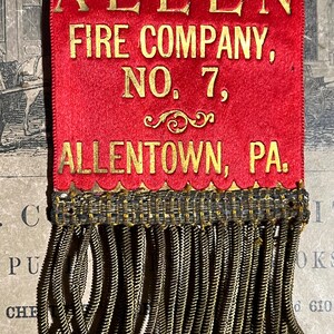 May include: A red ribbon with gold lettering that reads "ALLEN FIRE COMPANY, NO. 7, ALLENTOWN, PA." The ribbon has a decorative gold fringe at the bottom. The ribbon is likely a commemorative item.