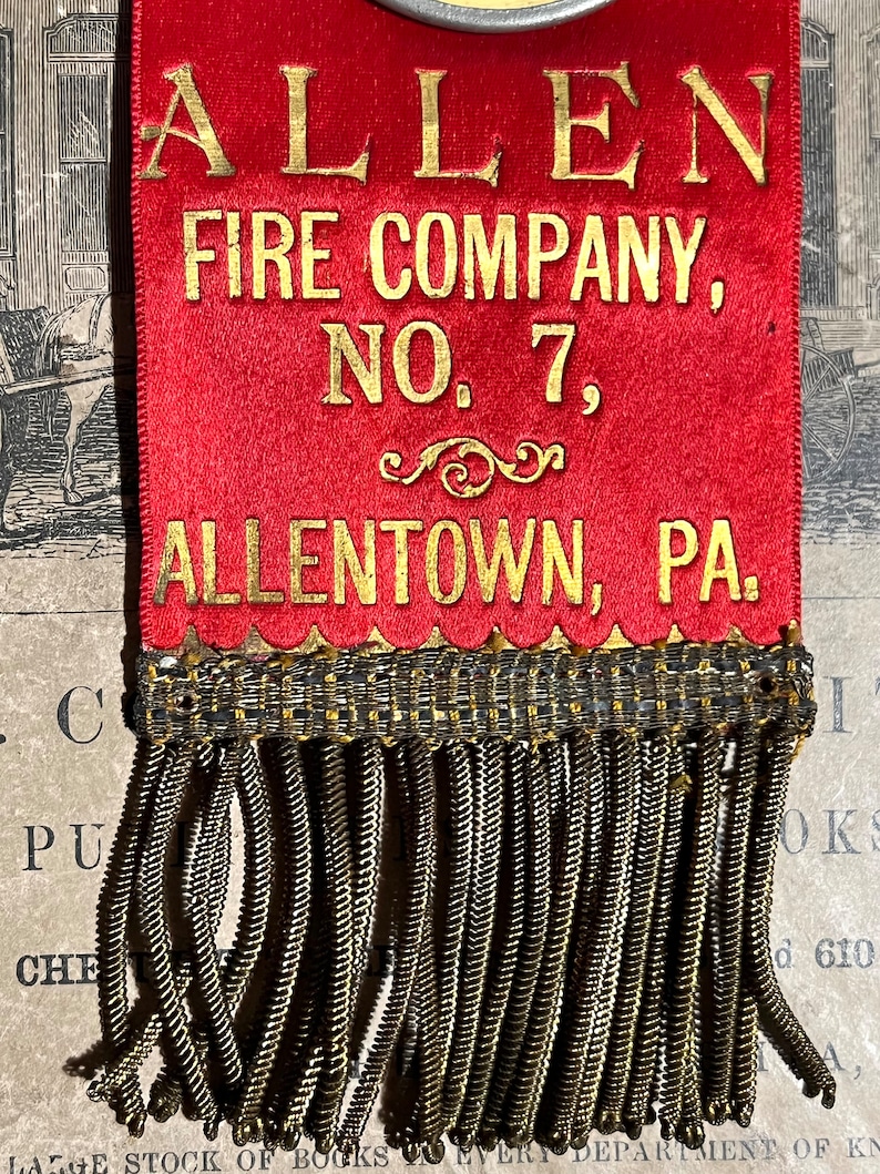 May include: A red ribbon with gold lettering that reads "ALLEN FIRE COMPANY, NO. 7, ALLENTOWN, PA." The ribbon has a decorative gold fringe at the bottom. The ribbon is likely a commemorative item.