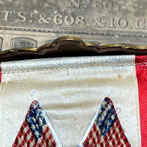 May include: A vintage banner with a decorative brass top. The banner features two embroidered American flags with red and white stripes and blue stars. The banner has a white background with red trim.