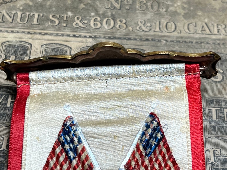 May include: A vintage banner with a decorative brass top. The banner features two embroidered American flags with red and white stripes and blue stars. The banner has a white background with red trim.