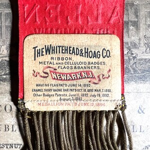 May include: A vintage red ribbon with embossed text and a rectangular card featuring the text "THE WHITEHEAD & HOAG CO." and other text. The card has a decorative fringe at the bottom. The ribbon is a historical artifact.