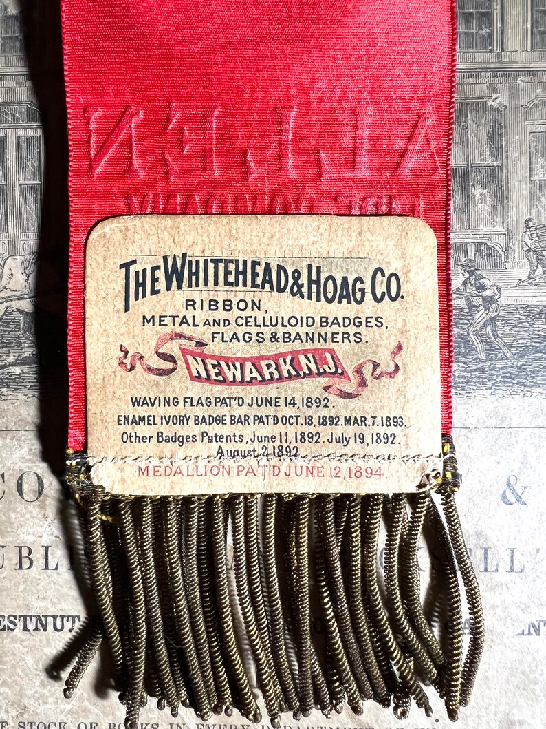 May include: A vintage red ribbon with embossed text and a rectangular card featuring the text "THE WHITEHEAD & HOAG CO." and other text. The card has a decorative fringe at the bottom. The ribbon is a historical artifact.
