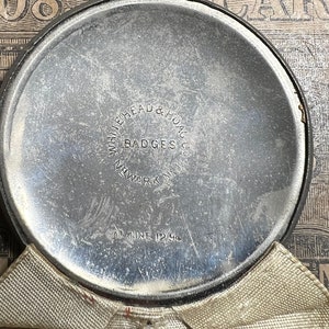May include: A circular, silver-toned metal object with the text "WHITEHEAD & HOAG CO. BADGES NEWARK N.J." and "AT JUNE 12 94" etched into the surface. The object is partially covered by a beige fabric.