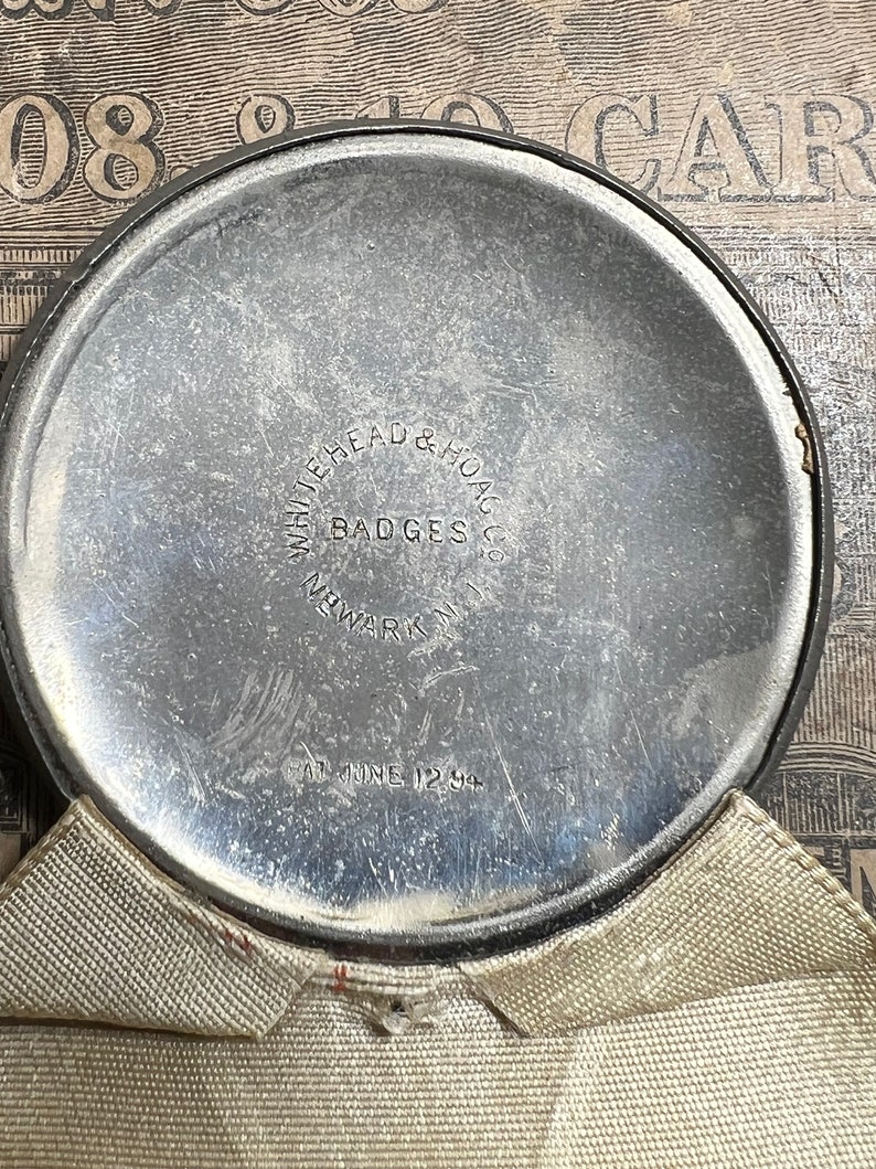 May include: A circular, silver-toned metal object with the text "WHITEHEAD & HOAG CO. BADGES NEWARK N.J." and "AT JUNE 12 94" etched into the surface. The object is partially covered by a beige fabric.