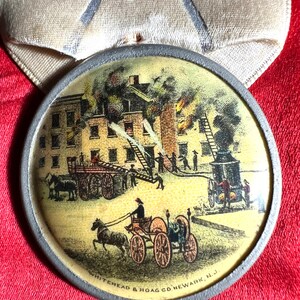 May include: A vintage round medal with a detailed illustration of a building fire scene. Firefighters are depicted using horse-drawn fire engines and ladders. The medal has a silver-colored rim and is set against a red background. The text "WHITEHEAD & HOAG CO. NEWARK, N.J." is visible.