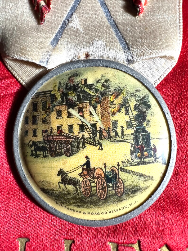 May include: A vintage round medal with a detailed illustration of a building fire scene. Firefighters are depicted using horse-drawn fire engines and ladders. The medal has a silver-colored rim and is set against a red background. The text "WHITEHEAD & HOAG CO. NEWARK, N.J." is visible.