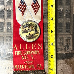 May include: Vintage fire company ribbon, approximately 9 inches long, featuring a firefighter illustration, crossed American flags, and a scene of a fire. The ribbon is red with gold lettering, and the bottom has a gold fringe. The text reads "ALLEN FIRE COMPANY, NO. 7, ALLENTOWN, PA."
