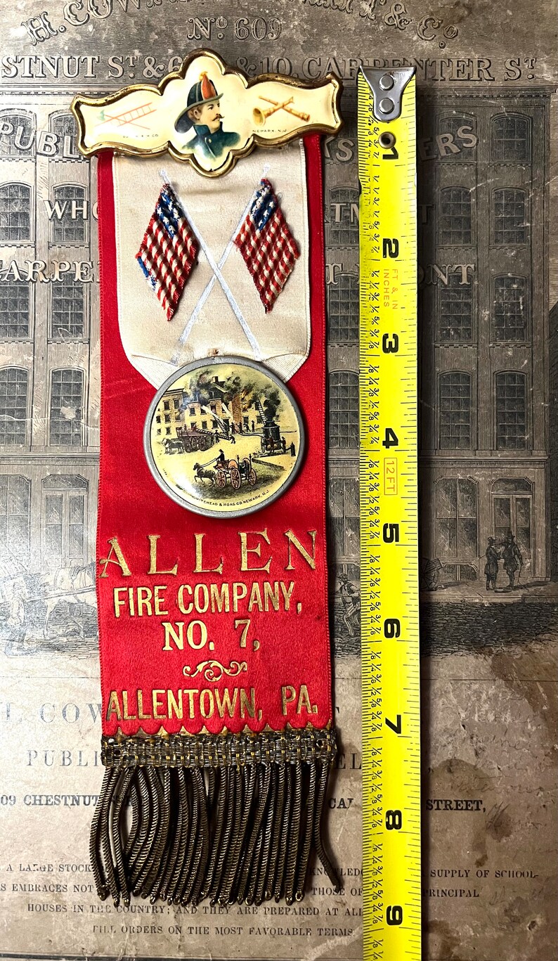 May include: Vintage fire company ribbon, approximately 9 inches long, featuring a firefighter illustration, crossed American flags, and a scene of a fire. The ribbon is red with gold lettering, and the bottom has a gold fringe. The text reads "ALLEN FIRE COMPANY, NO. 7, ALLENTOWN, PA."