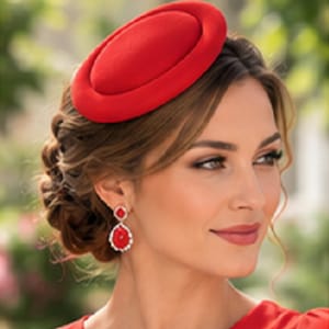 May include: A vibrant red pillbox hat with a smooth, rounded crown and a structured brim. The hat is worn at a slight angle on the head. Accompanying the hat are drop earrings featuring a central red stone surrounded by a border of small white crystals.