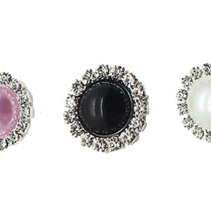 Puede incluir: Tres botones decorativos con una perla central en rosa, negro y blanco, cada uno rodeado por un anillo de brillantes pedrería. Los botones están sobre un fondo blanco.