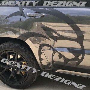 May include: A black and gray car with a large, stylized graphic of a snarling face on the side. The car has black rims and tires, with the text "GEXITY DEZIGNZ" in silver. The car door handle is visible.