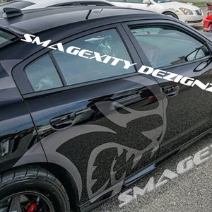 May include: Black car with white text that reads "SMAGEXITY DEZIGNZ". The car has a large grey graphic of a snarling face on the side. The car is parked on a paved surface.