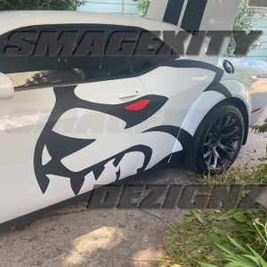 May include: White car with a black and red graphic design on the side. The graphic depicts a stylized animal head with sharp teeth and red eyes. The car has black wheels and a rear spoiler with black stripes. The text "SMAGEXITY DEZIGN" is visible.