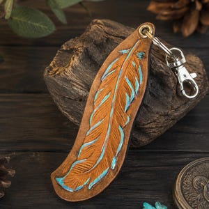 Puede incluir: Un llavero de cuero color canela con forma de pluma, con detalles en relieve y acentos en azul turquesa a lo largo de los bordes. La pieza de cuero está unida a un mosquetón giratorio de metal plateado y una anilla circular en la parte superior.