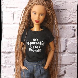 Könnte beinhalten: Eine Puppe mit langen braunen Zöpfen, die ein schwarzes T-Shirt mit dem Text "So Apparently I'm Dramatic" in Weiß trägt. Die Puppe trägt eine blaue Jeans und hat einen neutralen Gesichtsausdruck.