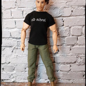 May include: A black t-shirt with the text "old school" in white lettering. The shirt is being worn by a doll with brown hair and green pants. The doll is standing in front of a white brick wall.