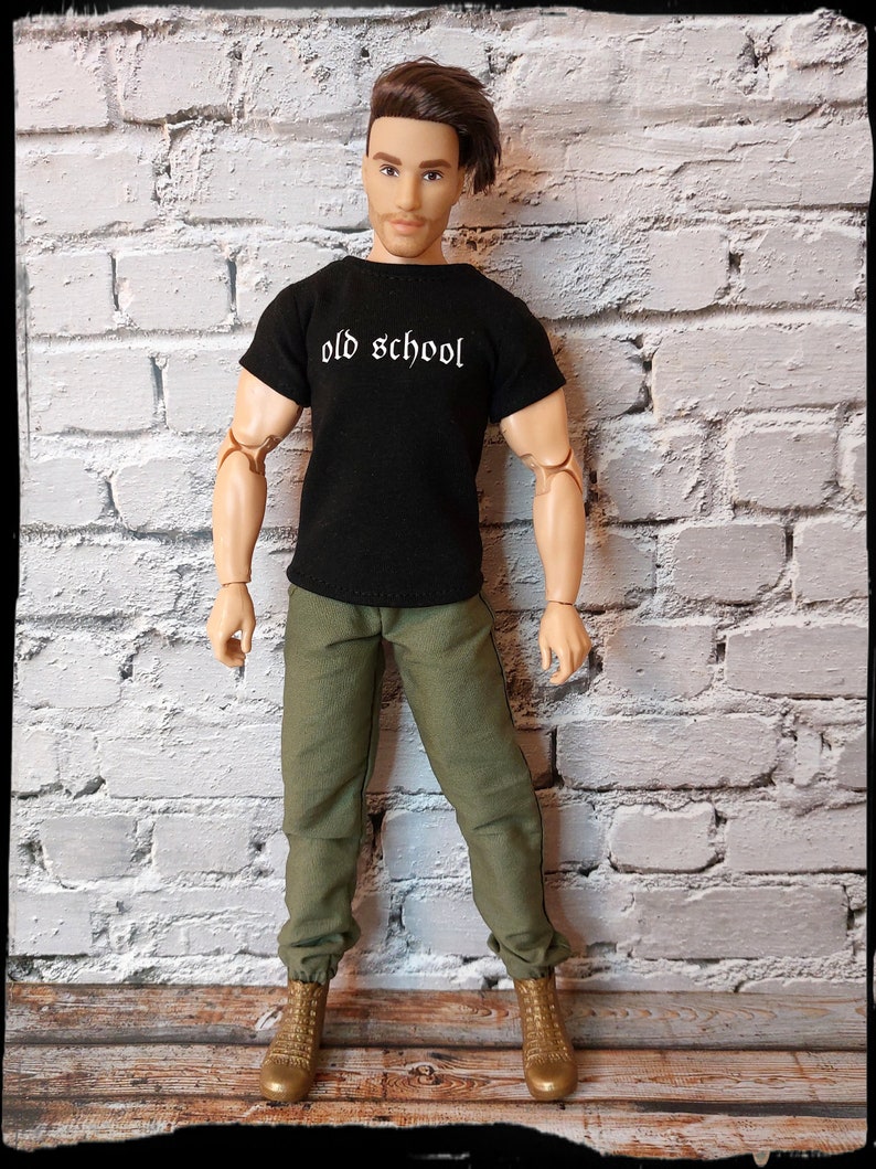 May include: A black t-shirt with the text "old school" in white lettering. The shirt is being worn by a doll with brown hair and green pants. The doll is standing in front of a white brick wall.