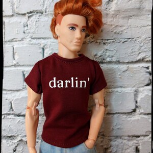 Pu&ograve; includere: Bambola con capelli rossi e occhi blu, indossa una maglietta bordeaux con la scritta "darlin'" in bianco e jeans blu. La bambola &egrave; posizionata contro un muro di mattoni bianchi.