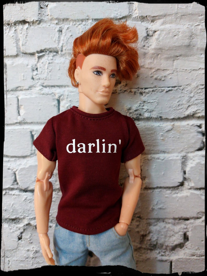 Pu&ograve; includere: Bambola con capelli rossi e occhi blu, indossa una maglietta bordeaux con la scritta "darlin'" in bianco e jeans blu. La bambola &egrave; posizionata contro un muro di mattoni bianchi.