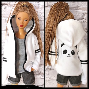 Peut inclure: Une poupée vêtue d'une veste à capuche blanche avec des rayures noires sur les manches et un visage de panda au dos. La poupée porte également un débardeur gris et un short en jean. La poupée a de longs cheveux bruns tressés.