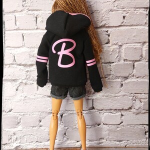 Pode incluir: Uma jaqueta de moletom com capuz preta com uma letra "B" rosa na frente. A jaqueta tem listras brancas nas mangas. A boneca est&aacute; usando shorts jeans e t&ecirc;nis brancos.