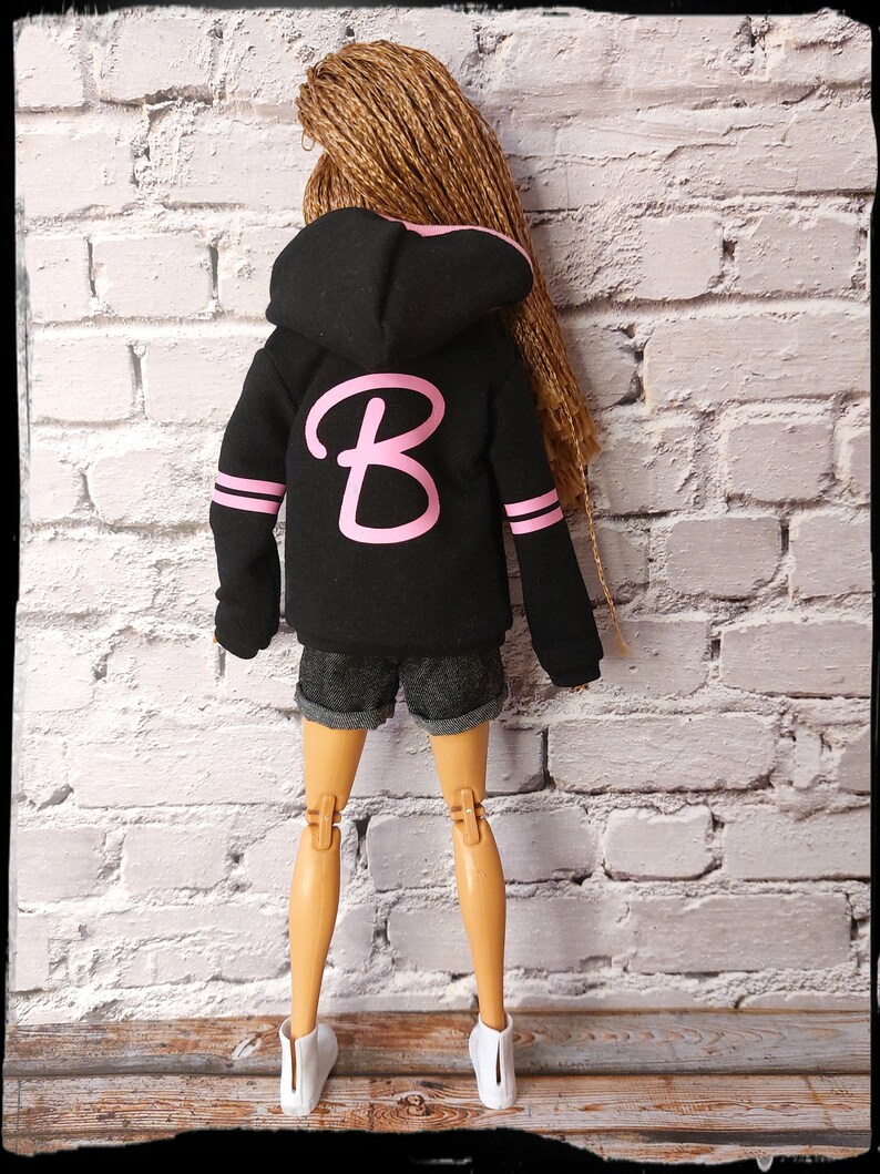 Pode incluir: Uma jaqueta de moletom com capuz preta com uma letra "B" rosa na frente. A jaqueta tem listras brancas nas mangas. A boneca est&aacute; usando shorts jeans e t&ecirc;nis brancos.